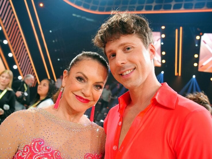 Christine Neubauer im glitzernden Kleid mit langen pinken Ohrringen und Valentin Lusin in offenem roten Hemd lächeln in die Kamera.