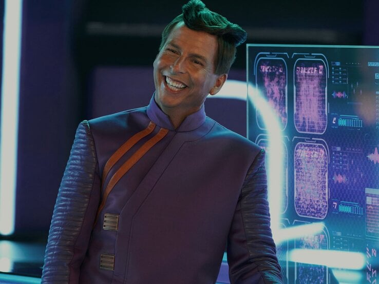 Jake McBrayer mit markantem, hochgestyltem Haar lächelt breit in einem futuristischen lila Anzug mit orangefarbenen Akzenten vor einem digitalen Bildschirm. | © Steve Wilkie/ Apple TV+
