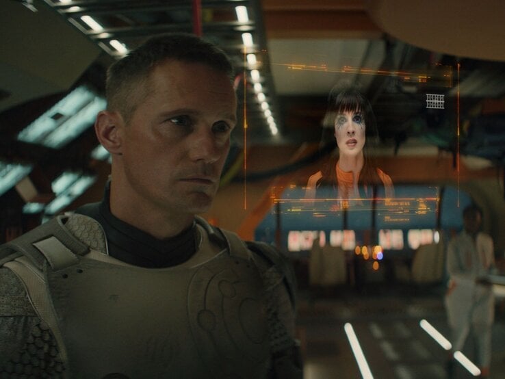 Alexander Skarsgård in einem futuristischen Raumanzug sieht nachdenklich auf ein holografisches Bild einer Frau mit markanten Make-up, das vor ihm schwebt. | © Steve Wilkie/ Apple TV+