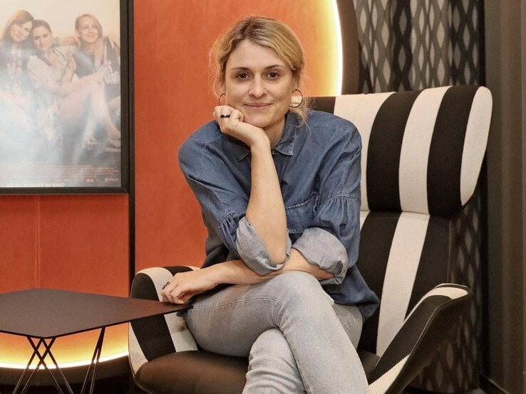 Julia Becker mit blonder Hochsteckfrisur sitzt lächelnd in Jeans und Jeanshemd auf einem schwarz-weiß gestreiften Sessel.