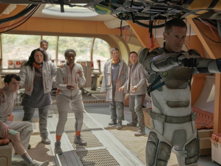 Alexander Skarsgard in "Murderbot": Ein ernster Mann in futuristischer Rüstung zielt mit erhobenen Armen, während sieben verängstigte Personen hinter ihm stehen.