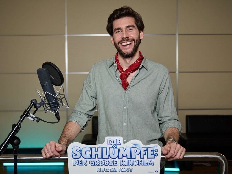 Alvaro Soler mit braunem Bart und grünem Hemd steht vor Mikrofon, während er für "Die Schlümpfe" Werbung macht.