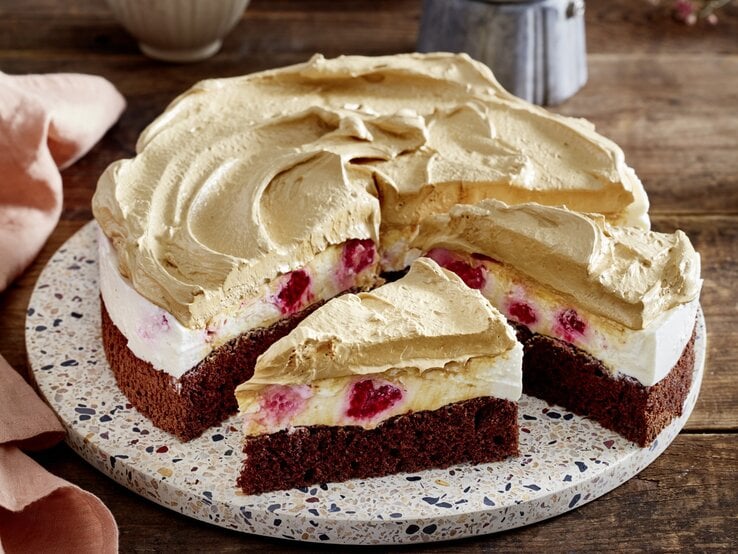 Dalgona-Himbeer-Cheesecake auf einem Tortenteller auf hölzernem Hintergrund. Links davon ein rosarotes Geschirrtuch.  | © EAT CLUB/Isabell Triemer