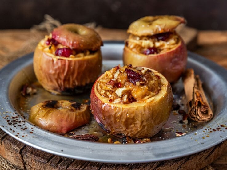 Drei ofengebackene Äpfel mit nussiger Füllung und Cranberries in einem Metallteller, dekoriert mit Zimtstangen auf rustikalem Untergrund. | © Shutterstock/Viktoria Hodos