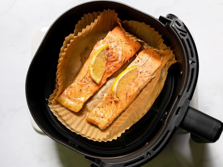 Zwei goldbraun gebackene Lachsfilets mit Zitronenscheiben und Gewürzen liegen auf Backpapier in einem schwarzen Airfryer. | © Shutterstock/PJjaruwan