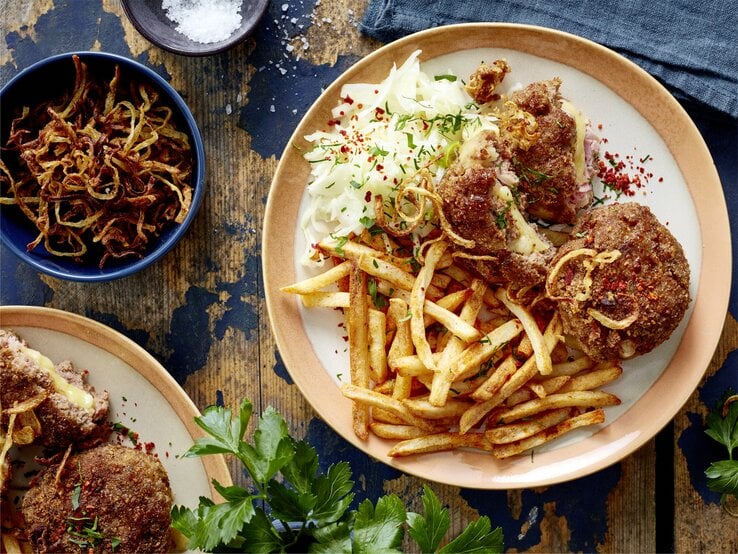  Cordon-bleu-Frikadellen mit Pommes auf einem Teller auf hölzernem Hintergrund. Links davon Röstzwiebeln, Salz und weitere Frikadellen.