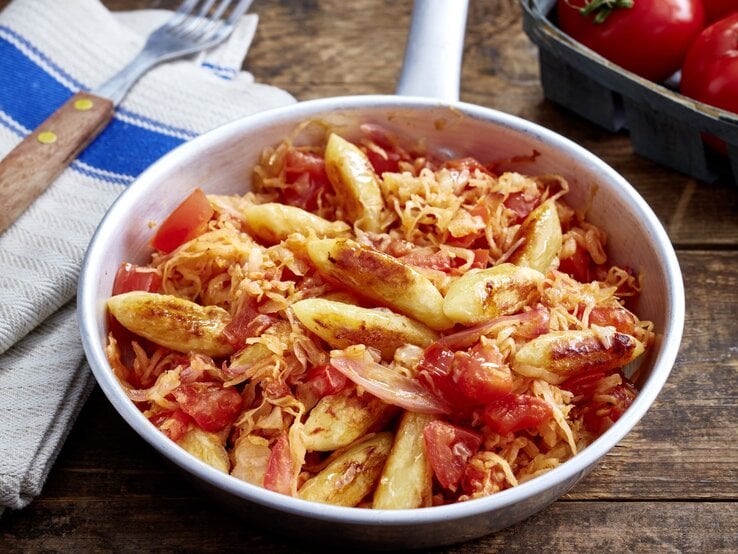 Schupfnudel-Sauerkraut-Pfanne auf hölzernem Hintergrund. Links Besteck und ein Geschirrtuch, oben auf dem Foto eine Tomate.