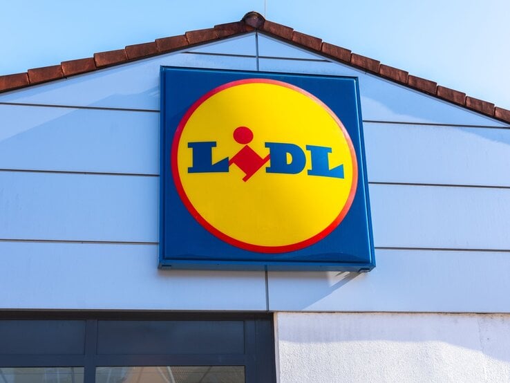 Lidl-Logo in gelber Kreisform mit rotem Rand an der hellblauen Fassade eines Filialgebäudes unter klarem Himmel.