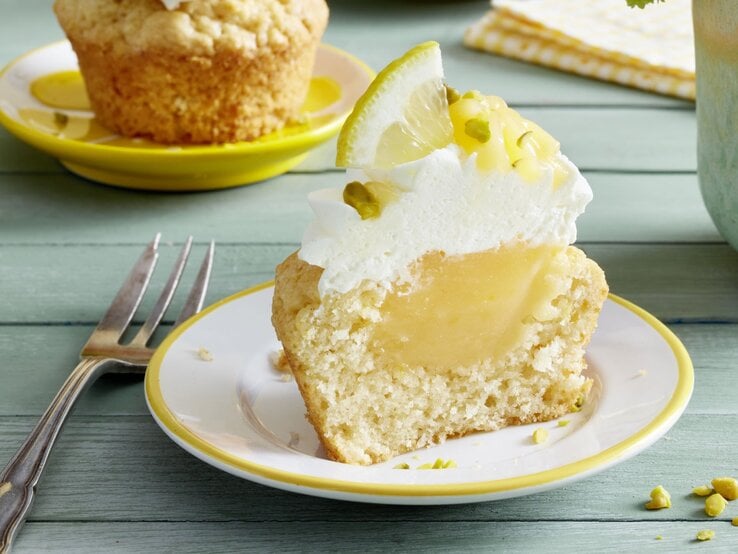 Zitronen-Cupcakes mit cremiger Füllung, Sahnetuff und Zitronenscheibe als Dekoration auf einem Teller, garniert mit gehackten Pistazien – im Hintergrund ein weiterer Cupcake und eine Gabel auf einem mintgrünen Holztisch.