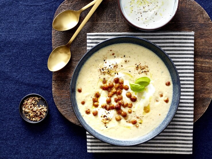 Cremiges Knoblauch-Blumenkohl-Süppchen in einer blauen Schale, getoppt mit knusprigen Kichererbsen, einem Klecks Joghurt, frischem Basilikum und Gewürzen, serviert auf einem Holzbrett mit gestreiftem Stoff und goldenen Löffeln daneben.