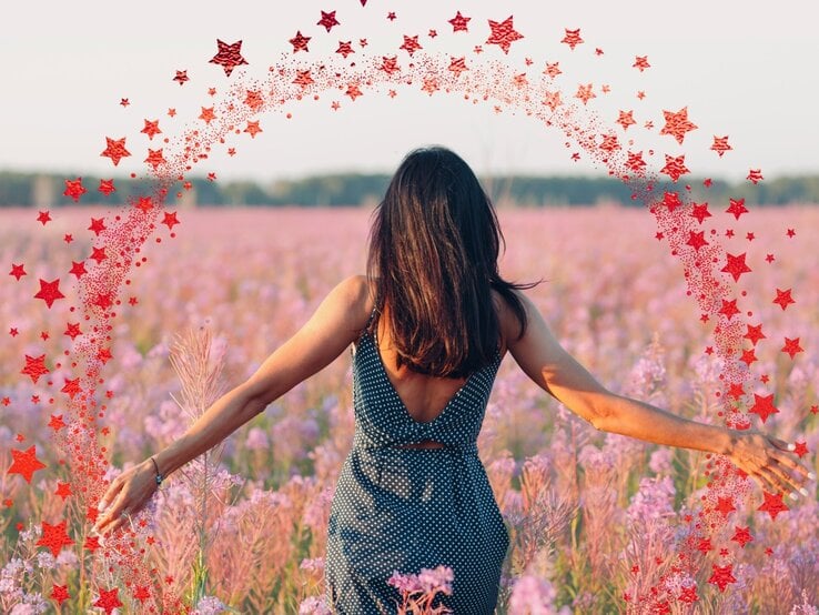 Eine Frau mit dunkelbraunem Haar in einem ärmellosen, gepunkteten Kleid steht mit dem Rücken zur Kamera in einem weiten Feld voller rosa-lila Wildblumen. Ihre Arme sind seitlich ausgestreckt, während sie die Blumen berührt. Um sie herum ist ein digital eingefügter Halbkreis aus roten Sternen zu sehen, der wie ein magischer Funkenbogen wirkt. Der Himmel ist hell und klar, im Hintergrund ist eine Baumlinie zu erkennen.