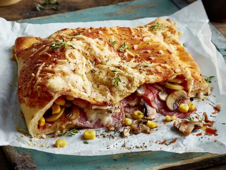 Pizza Calzone mit Mais, Pilzen und Salami auf weißem Backpapier vor hölzernem, leicht blau bemaltem Hintergrund.