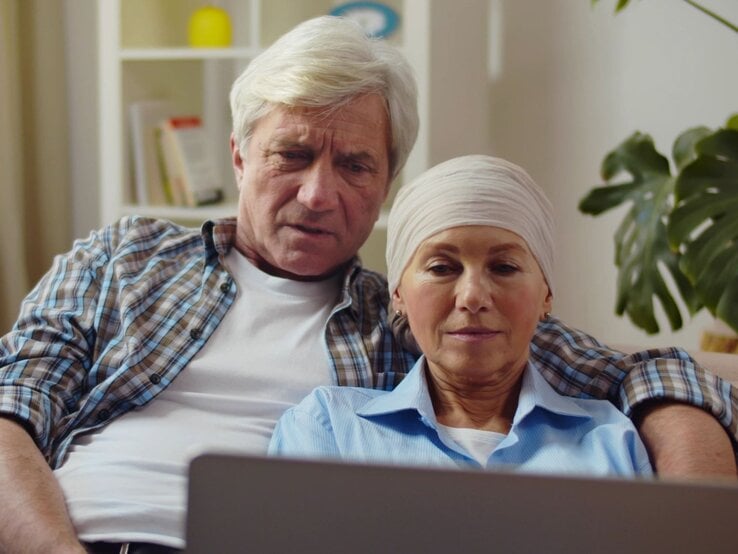 Nachdenkliches Paar in gemütlicher Umgebung, die Frau mit Brustkrebs wirkt ruhig und wird vom Partner gehalten.