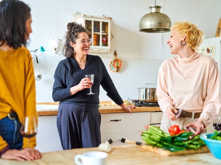 Drei Frauen stehen bei Vorbereitungen fürs Essen in der Küche. Sie unterhalten sich mit einem Glas Wein in der Hand und die Gemeinschaft macht ihnen Spaß.