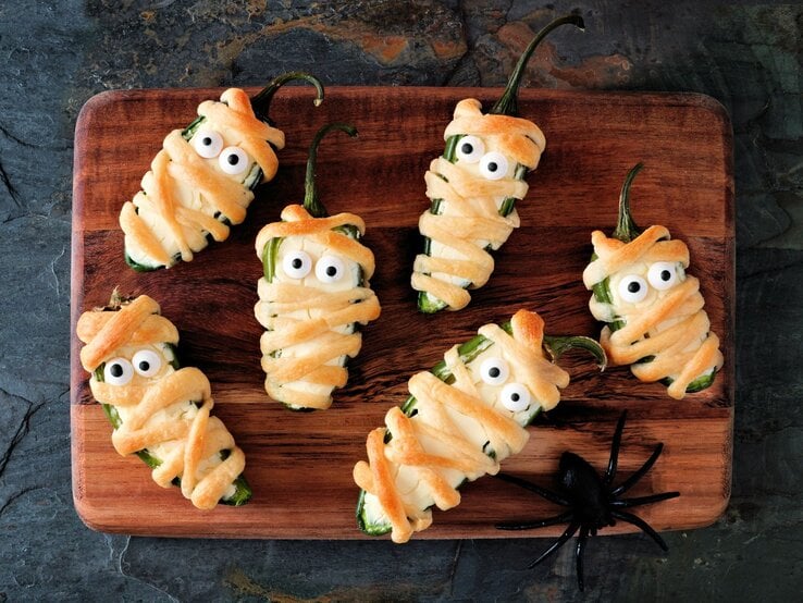 Sechs Mumien Jalapeno Popper auf einem Holzbrett – halbierte Jalapeños, mit Frischkäse gefüllt, in Teigstreifen gewickelt und mit Zuckeraugen dekoriert, als lustiger Halloween-Snack angerichtet.