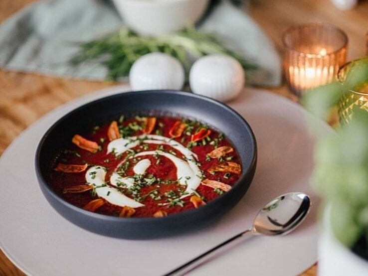 Eine Schale Rotkohl-Schmand-Suppe mit Karotten-Chips mit dekorativem Sahne-Wirbel, gehackten Kräutern und knusprigen Topping-Streifen, serviert auf einem gedeckten Tisch mit Kerzenlicht und einem Löffel daneben.