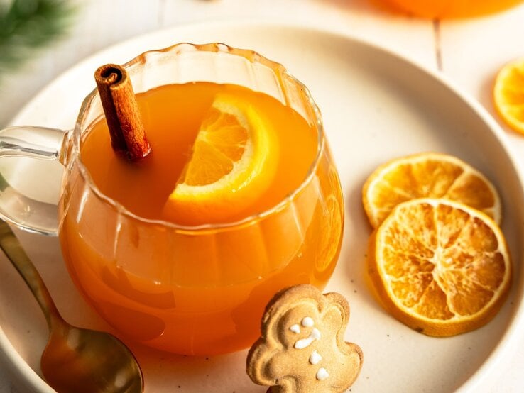Ein Glas mit alkoholfreier Orangenpunsch, dekoriert mit einer Orangenscheibe und einer Zimtstange, steht auf einem Teller. Neben dem Glas liegen getrocknete Orangenscheiben, ein goldener Löffel und ein Lebkuchenplätzchen in Form eines Lebkuchenmanns. Die warme und gemütliche Szene vermittelt Winter- und Weihnachtsstimmung.