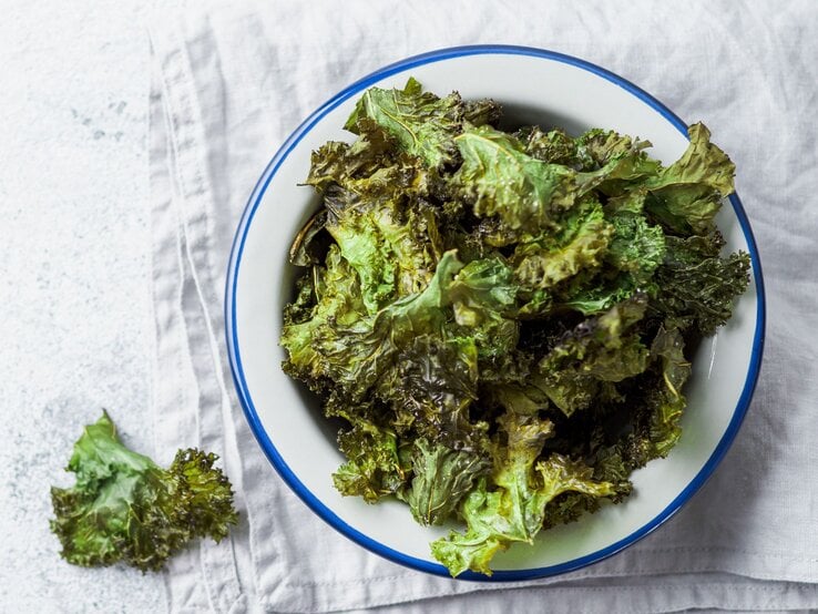 Draufsicht auf eine Schüssel mit knusprigen Grünkohl-Chips, daneben ein einzelnes Blatt auf einem hellen Untergrund, arrangiert auf einem Tuch.