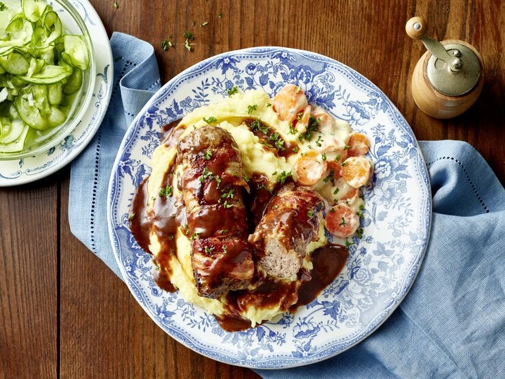 Draufsicht auf einen gut gefüllten Teller mit den kleinen Räuber-hackbraten, dem Kartoffelpüree und den gestovten Möhren auf einem rustikalen Untergrund.