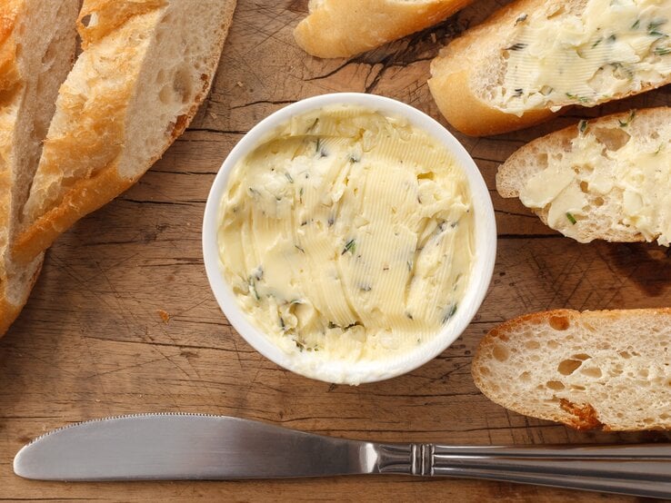 Butter mit Zitronenverbene in einer kleinen runden Form / Baguette ganz und aufgeschnitten / Streichmesser aus Metall / Untergrund grobes Holz