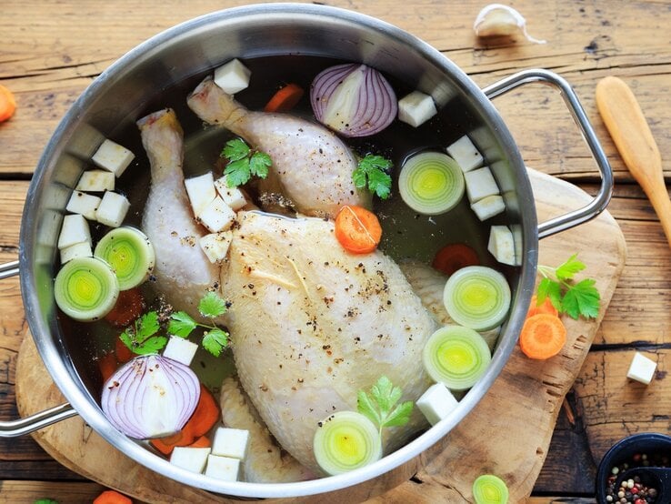 Hühnerbrühe im Topf / ganzen Suppenhuhn, klein geschnittenes Gemüse / Unterlagen Holzbrett auf Holzboden / frischer Pfeffer in Mörser / Holzlöffel / frisches geschnittenes Gemüse - Möhren, Knoblauch, Sellerie, Petersilie