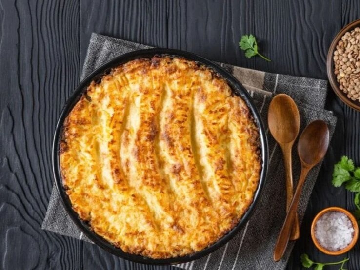 Veganer Linsen-Gemüse-Auflauf in runder Auflaufform auf dunklem Holztisch mit Linsen und Kräutern.